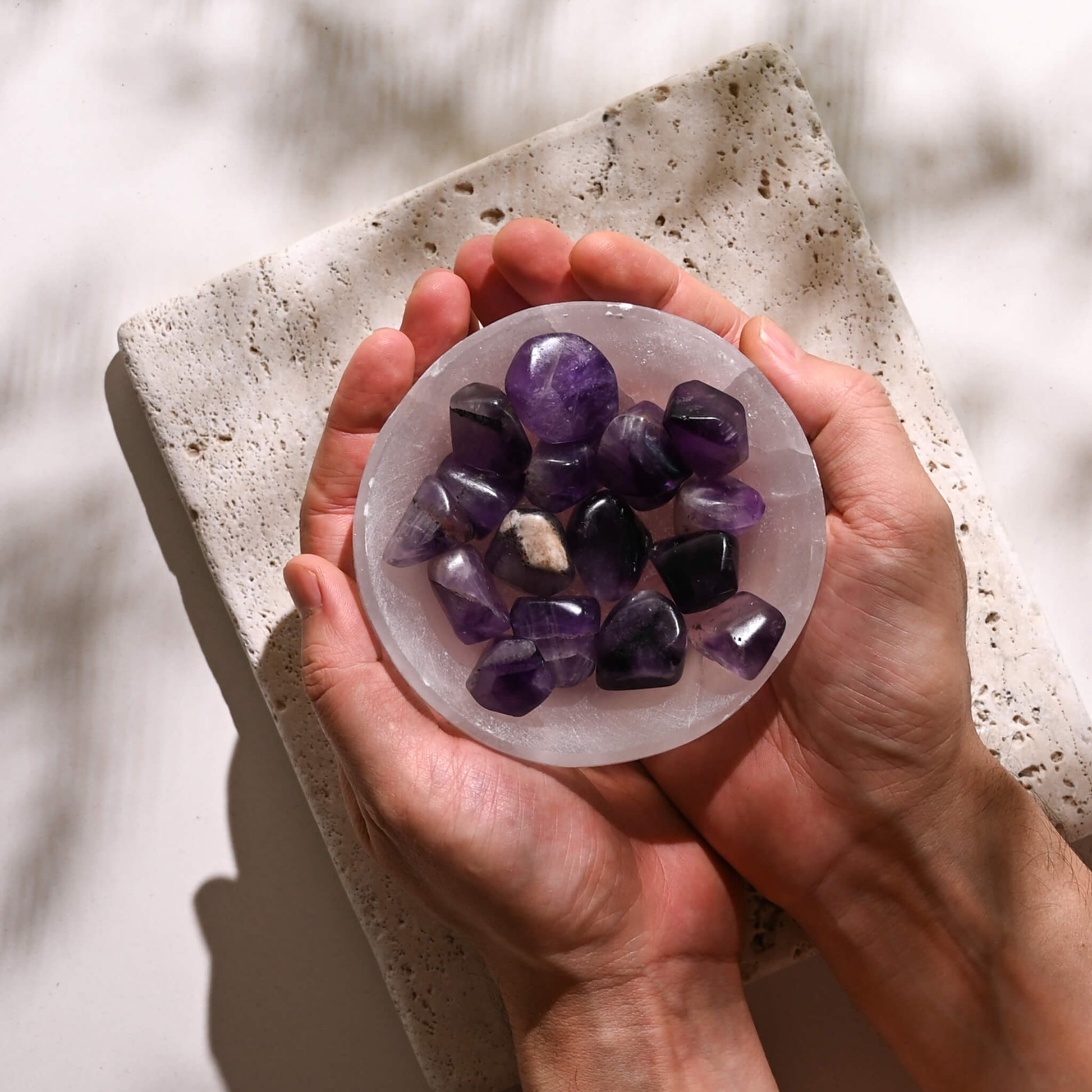 Hands holding a selenite bowl fillled with Amethyst Tumbled Stones